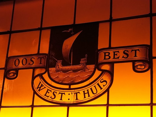 A stained glass window with a warm orange glow features an emblem of a ship with a sail. Surrounding the ship are banner scrolls with the words 'OOST WEST THUIS BEST', which roughly translates to 'East West Home Best'. The ship is set against a backdrop of stylized waves.