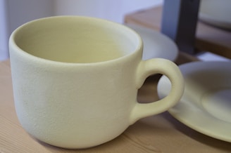A large, textured ceramic mug sits on a wooden surface next to a matching plate. The mug has a rough, matte finish with a thick handle and a simple, minimalist design.