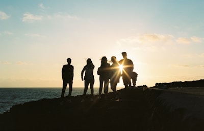 silhouette of people standing on rock formation near body of water during sunset