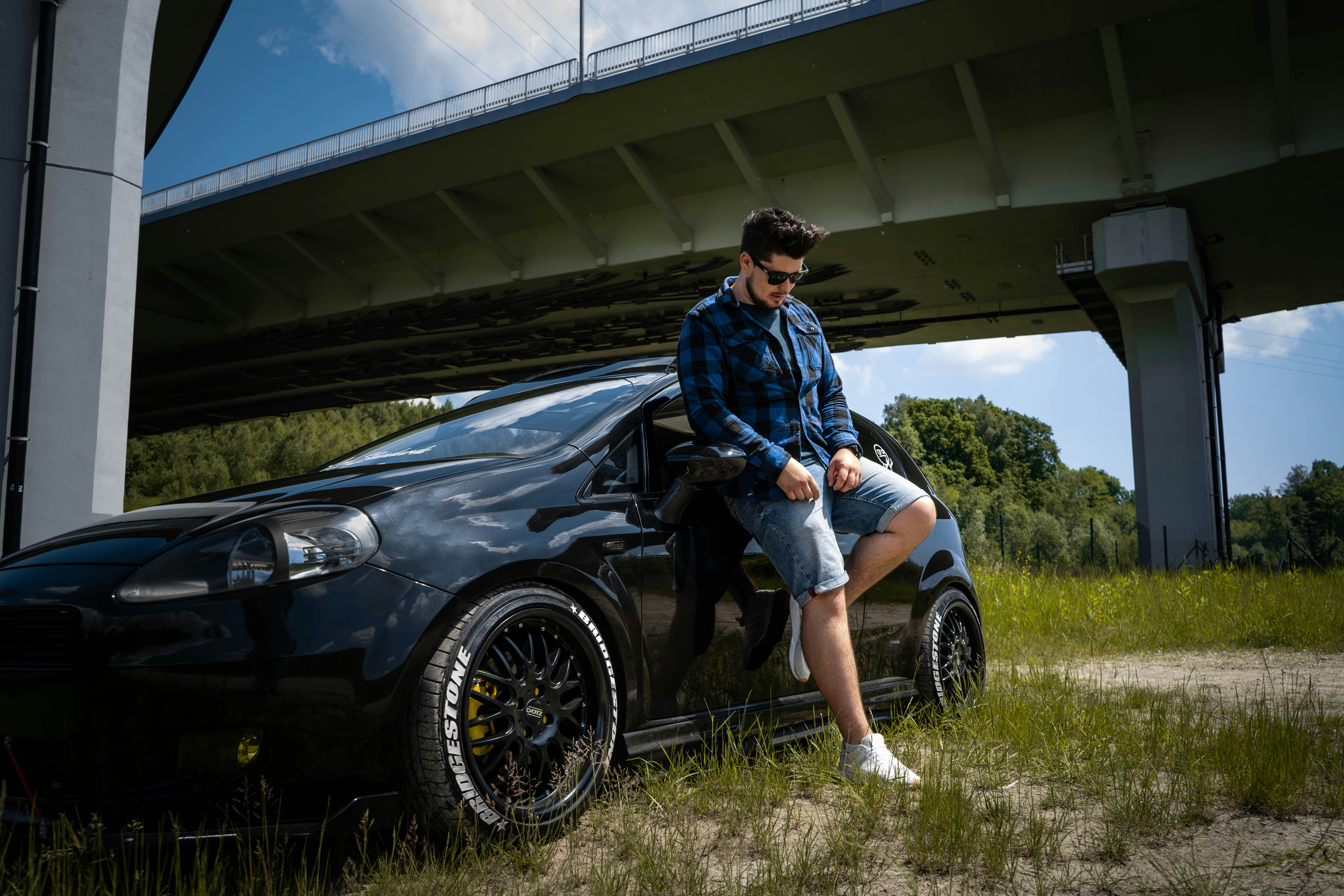 Femme en chemise à fleurs bleue et blanche et short en jean bleu s’appuyant sur une voiture noire