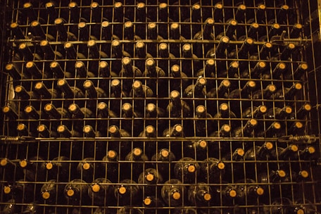 A neatly organized wine rack filled with rows of dark-colored wine bottles, each stacked on metal shelves. The lighting is dim, creating a warm and rustic atmosphere. The caps of the bottles are visible, indicating the bottles are stored horizontally.