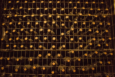 A neatly organized wine rack filled with rows of dark-colored wine bottles, each stacked on metal shelves. The lighting is dim, creating a warm and rustic atmosphere. The caps of the bottles are visible, indicating the bottles are stored horizontally.