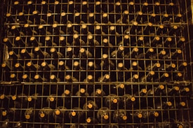 A neatly organized wine rack filled with rows of dark-colored wine bottles, each stacked on metal shelves. The lighting is dim, creating a warm and rustic atmosphere. The caps of the bottles are visible, indicating the bottles are stored horizontally.