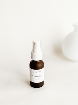 A small brown bottle with a white label and cap stands on a neutral background. The label reads 'ELDERFLOWER' and 'ROMI Apothecary Facial Toner Dry Skin'. A blurred white vase is positioned in the background, adding a minimalistic touch.