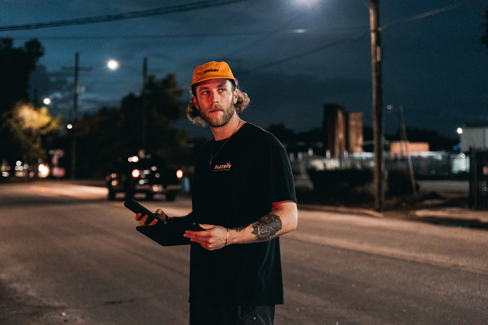 man in black crew neck t-shirt and orange cap holding black smartphone during nighttime