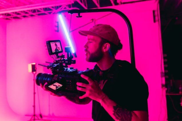 A person is operating a professional camera rig with a mounted monitor. The scene is illuminated with a strong pink light, creating a vibrant atmosphere. The individual is wearing a cap and has tattoos on their arm. The background is industrial with visible ceiling beams.