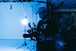 A person stands in a dimly lit studio holding a large professional camera, with bright white studio lights in the background. The setting has a moody, industrial feel, with visible equipment and a partially completed ceiling.