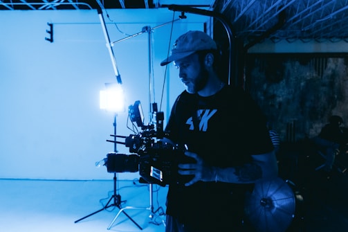 A person stands in a dimly lit studio holding a large professional camera, with bright white studio lights in the background. The setting has a moody, industrial feel, with visible equipment and a partially completed ceiling.
