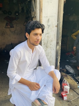 A person in traditional attire is sitting cross-legged in a rustic setting. The background shows a cluttered space with items such as cables, containers, and tools. The individual looks thoughtful and relaxed.