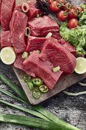 Slices of raw red meat are placed on a wooden cutting board alongside fresh vegetables. There are green chili slices, lemon halves, cherry tomatoes on vine, and lettuce. The background includes a rustic textured surface.