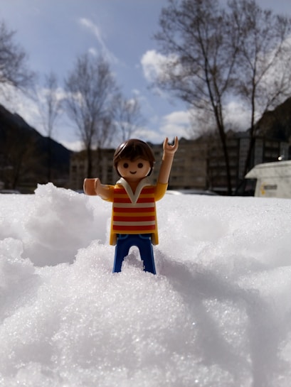A plastic toy figure with wide, smiling eyes is standing in a snowy landscape, dressed in a striped shirt and blue pants. The background features leafless trees and distant mountains under a partly cloudy sky.