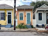 Three brightly colored houses are situated side by side on a residential street. Each house has a distinct color scheme: yellow, peach, and light gray. The houses feature shutters, steps leading to front doors, and are decorated with plants and a wreath. An American flag is displayed in front of the middle house. A utility pole and various signs are visible along the sidewalk, while chairs and a small table are placed in front of the gray house.