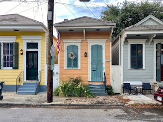 Three brightly colored houses are situated side by side on a residential street. Each house has a distinct color scheme: yellow, peach, and light gray. The houses feature shutters, steps leading to front doors, and are decorated with plants and a wreath. An American flag is displayed in front of the middle house. A utility pole and various signs are visible along the sidewalk, while chairs and a small table are placed in front of the gray house.