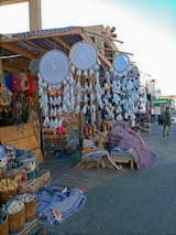A bazaar scene featuring a variety of handmade items such as large dreamcatchers with intricate designs and feather decorations. Shelves and tables display colorful textiles, woven baskets, and an assortment of crafts and souvenirs. The market stall has a canopy adorned with striped fabric, and there's a mixture of vibrant colors throughout the setup.