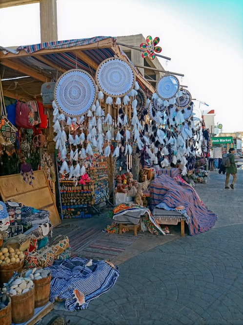 A bazaar scene featuring a variety of handmade items such as large dreamcatchers with intricate designs and feather decorations. Shelves and tables display colorful textiles, woven baskets, and an assortment of crafts and souvenirs. The market stall has a canopy adorned with striped fabric, and there's a mixture of vibrant colors throughout the setup.