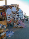 A bazaar scene featuring a variety of handmade items such as large dreamcatchers with intricate designs and feather decorations. Shelves and tables display colorful textiles, woven baskets, and an assortment of crafts and souvenirs. The market stall has a canopy adorned with striped fabric, and there's a mixture of vibrant colors throughout the setup.