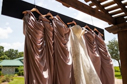 A collection of elegant dresses hanging on wooden hangers under a pergola, with a white lace bridal gown among several mauve bridesmaid dresses. The backdrop is a sunny outdoor setting with a green roofed building and lush greenery.