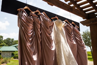 A collection of elegant dresses hanging on wooden hangers under a pergola, with a white lace bridal gown among several mauve bridesmaid dresses. The backdrop is a sunny outdoor setting with a green roofed building and lush greenery.
