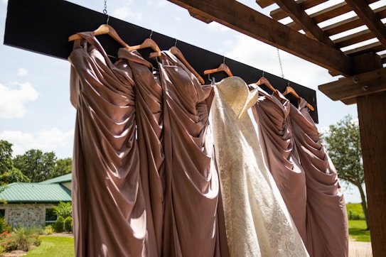 A collection of elegant dresses hanging on wooden hangers under a pergola, with a white lace bridal gown among several mauve bridesmaid dresses. The backdrop is a sunny outdoor setting with a green roofed building and lush greenery.