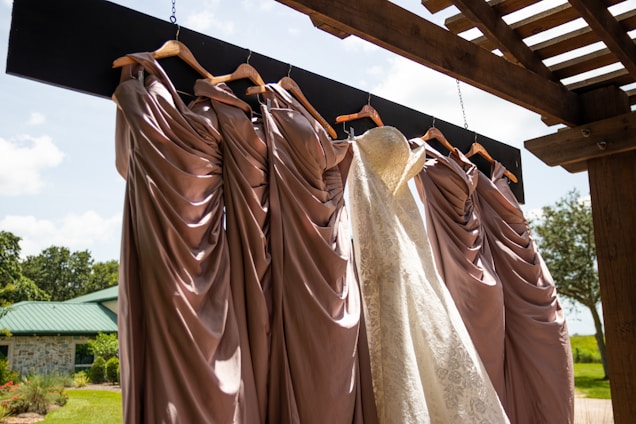 A collection of elegant dresses hanging on wooden hangers under a pergola, with a white lace bridal gown among several mauve bridesmaid dresses. The backdrop is a sunny outdoor setting with a green roofed building and lush greenery.