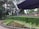 A well-maintained garden with neatly arranged flower beds featuring vibrant green bushes and various colored blooms. The scene is framed by a backdrop of tall, lush trees. To the right, a modern, angular structure with a dark, overhanging roof creates a contrast with the natural surroundings.