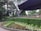 A well-maintained garden with neatly arranged flower beds featuring vibrant green bushes and various colored blooms. The scene is framed by a backdrop of tall, lush trees. To the right, a modern, angular structure with a dark, overhanging roof creates a contrast with the natural surroundings.