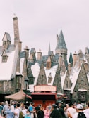 A bustling crowd gathers in front of a Butterbeer stand with snow-topped, medieval-style buildings in the background, featuring gabled roofs and stone chimneys. These structures evoke a fantastical, wizarding village scene.