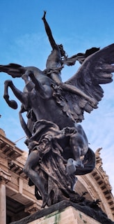 A dramatic bronze statue of a winged horse with a human figure riding it is prominently displayed against a clear blue sky. The sculpture is detailed and dynamic, capturing a sense of movement and mythology.