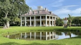 A large, elegant, colonial-style mansion with a symmetrical facade and large white columns sits amidst a well-manicured lawn. There is a small pond in the foreground reflecting the building, and lush green trees surround the area. The mansion has a steep, sloped roof with multiple dormer windows.