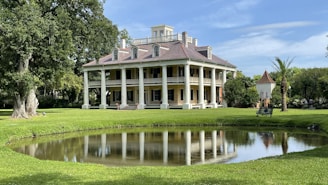 A large, elegant, colonial-style mansion with a symmetrical facade and large white columns sits amidst a well-manicured lawn. There is a small pond in the foreground reflecting the building, and lush green trees surround the area. The mansion has a steep, sloped roof with multiple dormer windows.