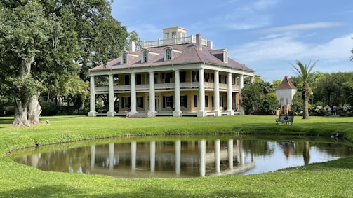 A large, elegant, colonial-style mansion with a symmetrical facade and large white columns sits amidst a well-manicured lawn. There is a small pond in the foreground reflecting the building, and lush green trees surround the area. The mansion has a steep, sloped roof with multiple dormer windows.