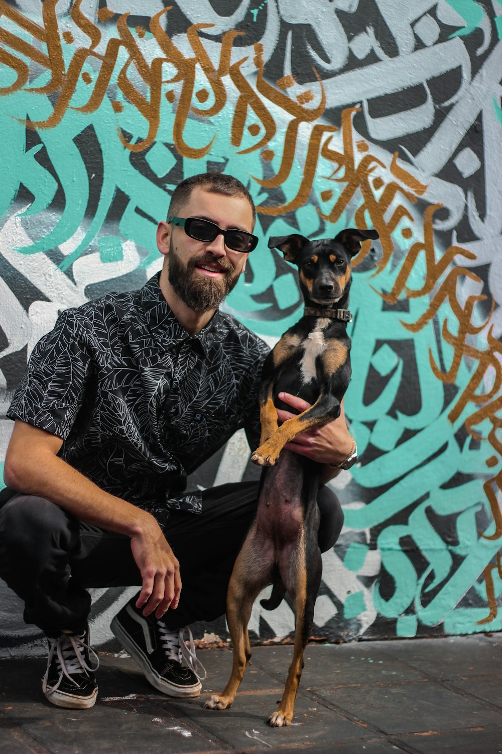 man in black and white floral shirt wearing black sunglasses holding brown short coated dog