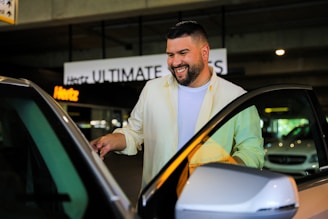 A man with a beard and a bright smile is standing by a car door, appearing to be opening or closing it. He is wearing a white shirt and a beige jacket. The background shows a parking area with signs for a rental car company, and other vehicles are visible.
