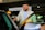 A man with a beard and a bright smile is standing by a car door, appearing to be opening or closing it. He is wearing a white shirt and a beige jacket. The background shows a parking area with signs for a rental car company, and other vehicles are visible.
