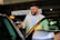 A man with a beard and a bright smile is standing by a car door, appearing to be opening or closing it. He is wearing a white shirt and a beige jacket. The background shows a parking area with signs for a rental car company, and other vehicles are visible.