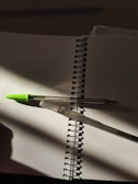 A spiral notebook lying open with blank pages, containing two pens intersecting over the pages. The pens are casting shadows. One pen has a green cap while the other is transparent. The lighting creates a contrast with areas of shadow and light.