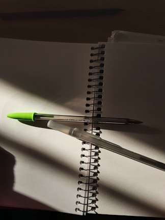A spiral notebook lying open with blank pages, containing two pens intersecting over the pages. The pens are casting shadows. One pen has a green cap while the other is transparent. The lighting creates a contrast with areas of shadow and light.