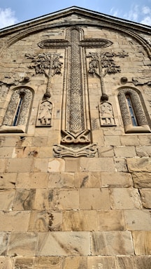 A stone facade featuring intricate carvings, including a large, ornate cross at the center surrounded by decorative elements and two human figures below. The architecture reflects a historical and religious design with a textured surface and aged appearance.