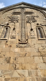 A stone facade featuring intricate carvings, including a large, ornate cross at the center surrounded by decorative elements and two human figures below. The architecture reflects a historical and religious design with a textured surface and aged appearance.