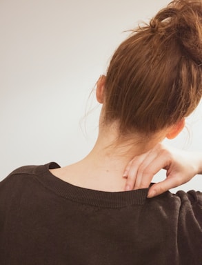 The image depicts the back of a person's head and shoulders, with the individual having brown hair tied up in a messy bun. A hand, with fingers gently curled, is placed on the neck, suggesting a sense of relaxation or relief. The individual is wearing a dark, soft-textured sweater.