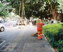 A person in an orange uniform and straw hat is cleaning a sidewalk with a broom. The walkway is lined with lush green bushes and large trees provide shade. Vehicles are parked by the roadside. Construction workers are visible further down the walkway with signage placed nearby, indicating an ongoing operation.