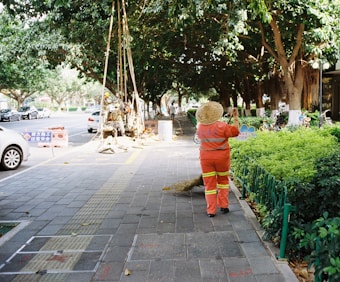 A person in an orange uniform and straw hat is cleaning a sidewalk with a broom. The walkway is lined with lush green bushes and large trees provide shade. Vehicles are parked by the roadside. Construction workers are visible further down the walkway with signage placed nearby, indicating an ongoing operation.