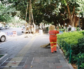 A person in an orange uniform and straw hat is cleaning a sidewalk with a broom. The walkway is lined with lush green bushes and large trees provide shade. Vehicles are parked by the roadside. Construction workers are visible further down the walkway with signage placed nearby, indicating an ongoing operation.