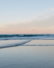 A serene ocean view with gentle waves lapping against the shoreline. The sky is a soft blend of pastel colors with hints of pink and blue. The calm water reflects the sky, creating a tranquil atmosphere.