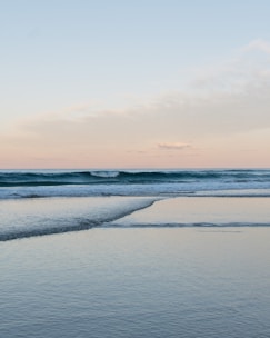 A serene ocean view with gentle waves lapping against the shoreline. The sky is a soft blend of pastel colors with hints of pink and blue. The calm water reflects the sky, creating a tranquil atmosphere.