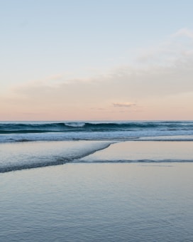 A serene ocean view with gentle waves lapping against the shoreline. The sky is a soft blend of pastel colors with hints of pink and blue. The calm water reflects the sky, creating a tranquil atmosphere.