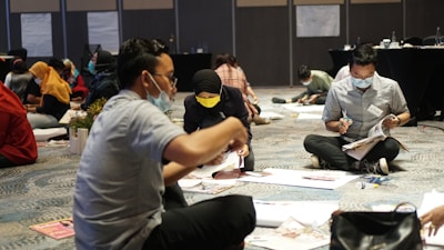 A group of people sitting on the floor, wearing face masks and engaged in an activity involving magazines and paper. The participants appear to be cutting or working on some form of project, possibly in a workshop or team-building setting. The setting is indoor, with a carpeted floor and conference tables in the background.