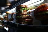 A row of stacked sandwiches filled with fresh greens, cheese, and meats all wrapped in white paper sits on a reflective surface under bright lighting.