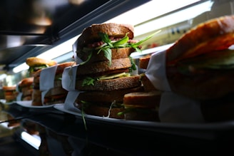 A row of stacked sandwiches filled with fresh greens, cheese, and meats all wrapped in white paper sits on a reflective surface under bright lighting.