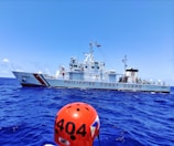 A Philippine Coast Guard vessel sails on a vibrant blue ocean. The ship is white with red stripes and the words 'PHILIPPINE COAST GUARD' are prominently displayed. A figure wearing an orange helmet labeled '4404' is visible in the foreground.